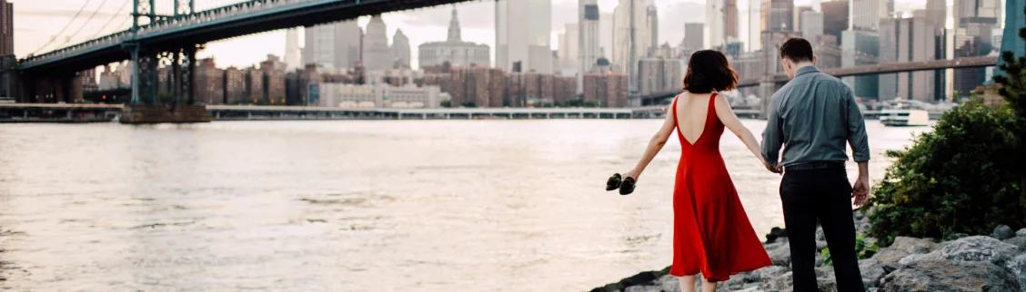 Elegant couple holding hands by waterfront with Manhattan Bridge and NYC skyline at golden hour
